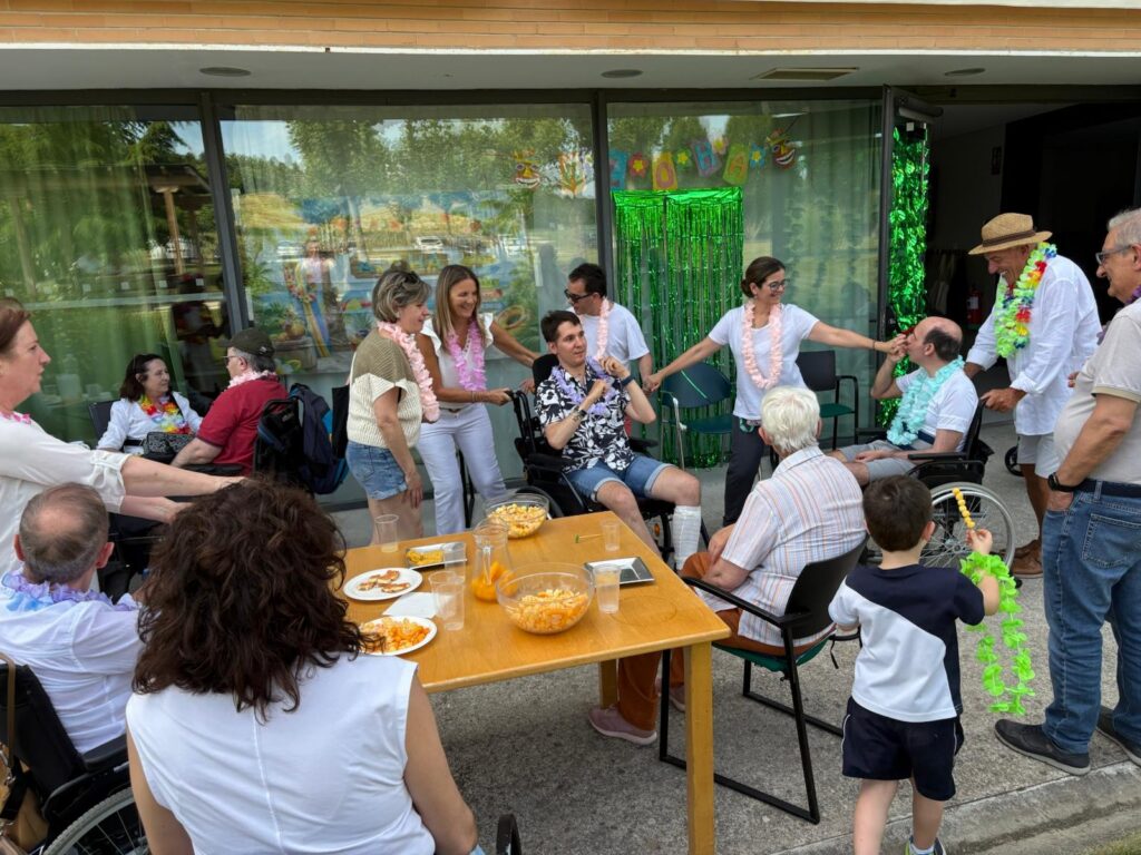 En la foto se ve a las personas usuarias de la residencia de Adacen junto con profesionales de la asociación y familiares en el porche de la residencia. Van vestidos de blanco con collares hawaianos