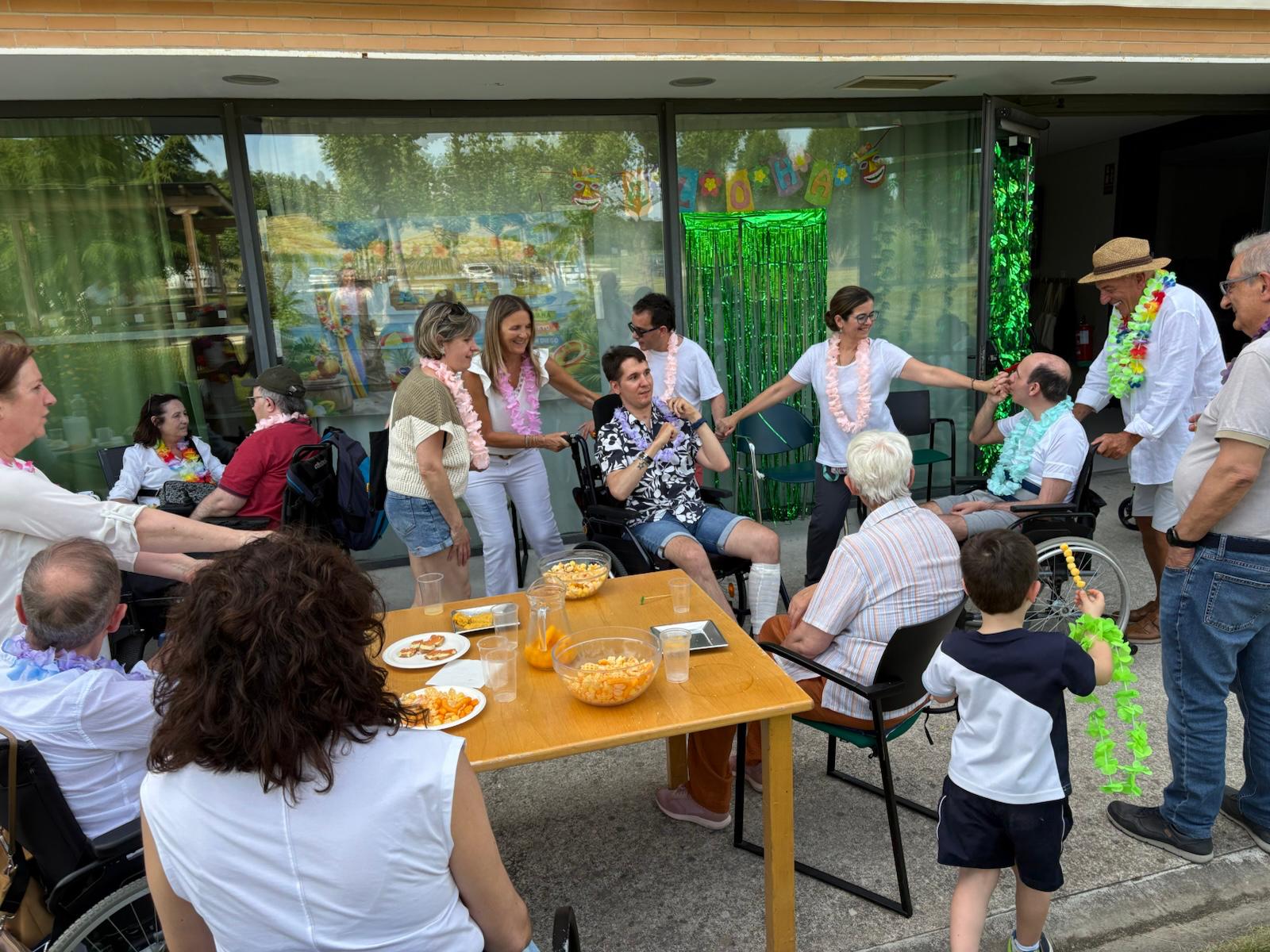 En la foto se ve a las personas usuarias de la residencia de Adacen junto con profesionales de la asociación y familiares en el porche de la residencia. Van vestidos de blanco con collares hawaianos