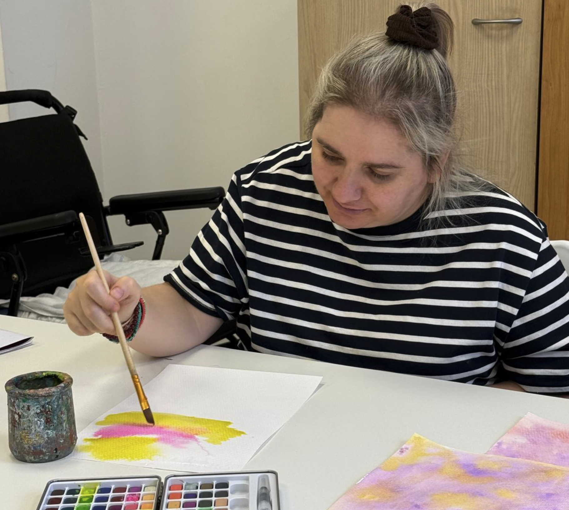 En la imagen se ve a una mujer sentada ante una mesa pintando con acuarela en colores rosa y amarillo