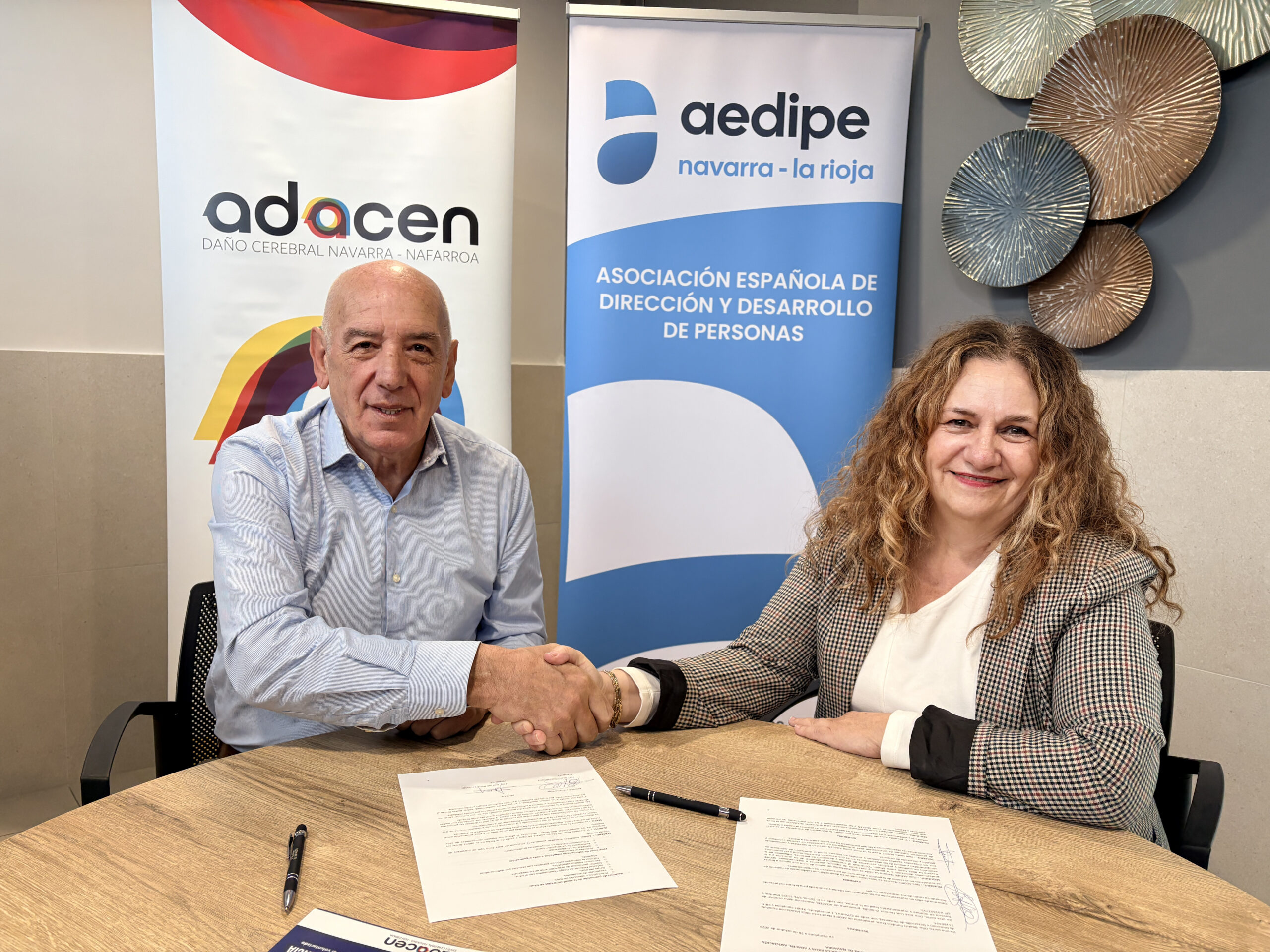 En la imagen se ve al presidente de Adacen y la Presidenta de AEDIPE sentados frente a una mesa dándose la mano tras firmar el convenio, que tienen delante